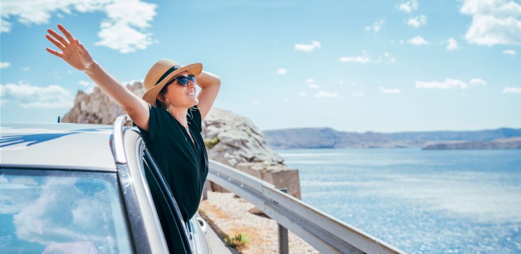 Glückliche Frau genießt ihren Urlaub, lehnt sich aus einem Autofenster, während sie eine malerische Küstenstraße entlangfährt, mit blauem Himmel und Meer im Hintergrund