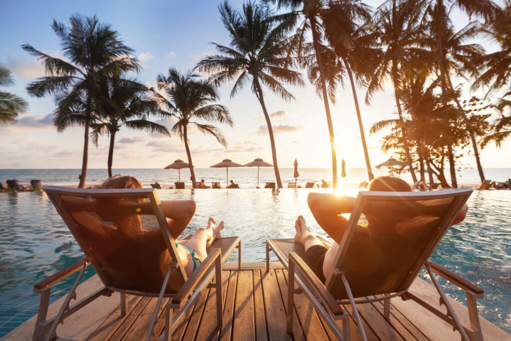 Zwei Personen entspannen auf Liegestühlen am Pool, umgeben von Palmen, mit Blick auf das Meer bei Sonnenuntergang.