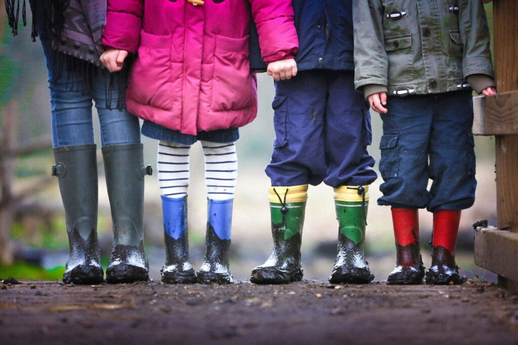 Vier Kinder in farbigen Gummistiefeln, die draußen im Matsch stehen, symbolisieren spielerisches Lernen im Freien.