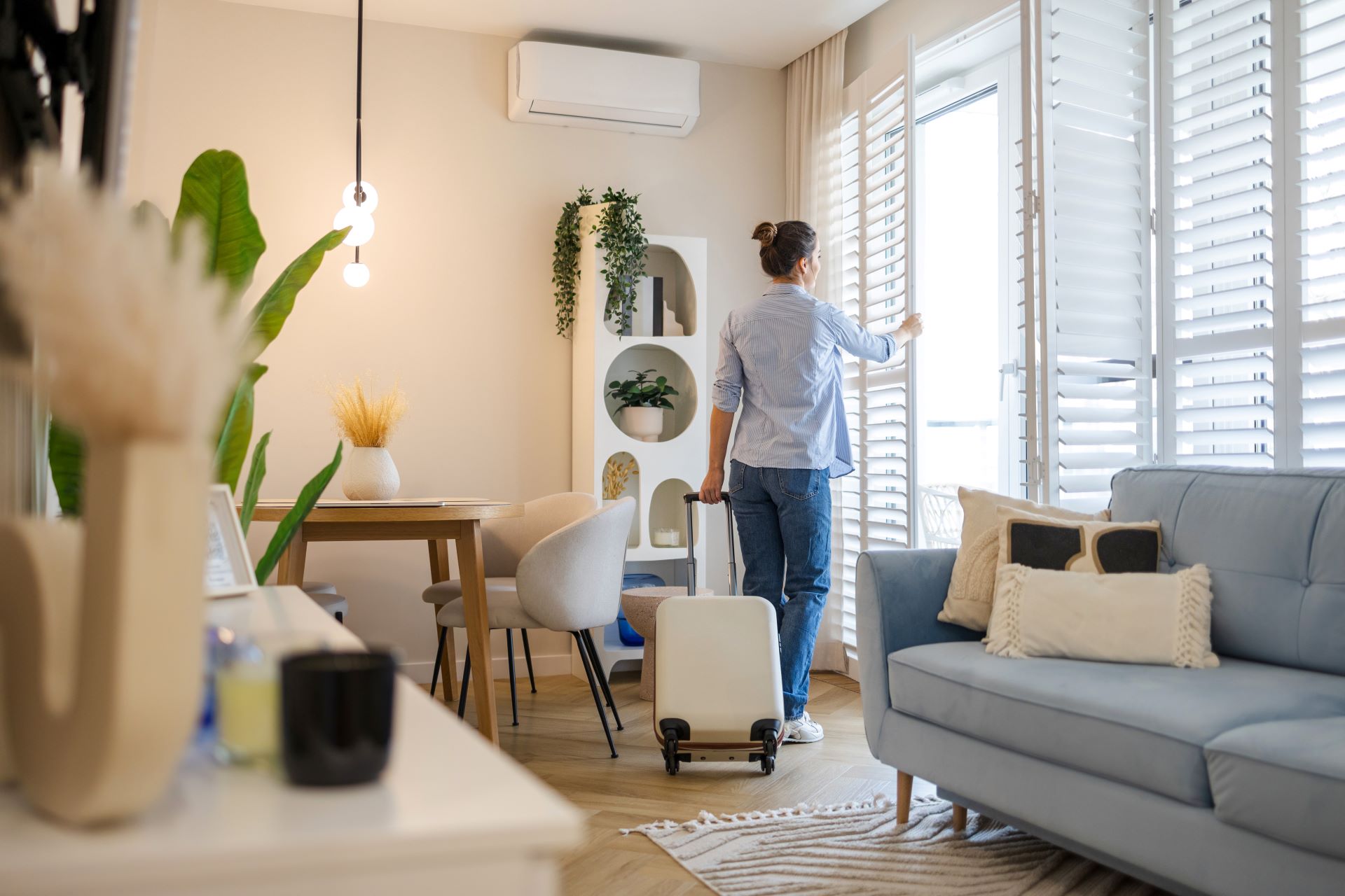 Moderne Ferienwohnung mit heller Einrichtung – eine Frau mit Koffer öffnet die Fensterläden und genießt die Aussicht.